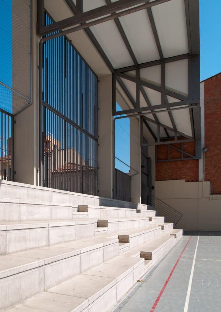 Detall constructiu: grades de formigó i coberta metàl·lica en pati escolar.