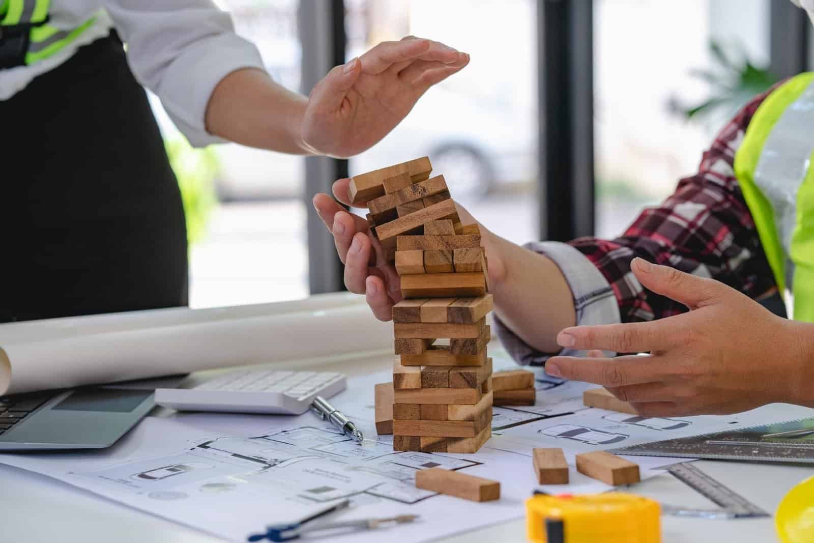 Técnicos revisando planos de obra mientras sostienen una torre inestable de bloques de madera sobre una mesa de trabajo.